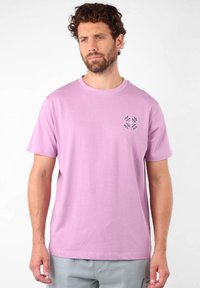 T-shirt en coton rose clair avec un col rond et des manches courtes. Présente un petit design décoratif avec des feuilles vertes sur la poitrine.