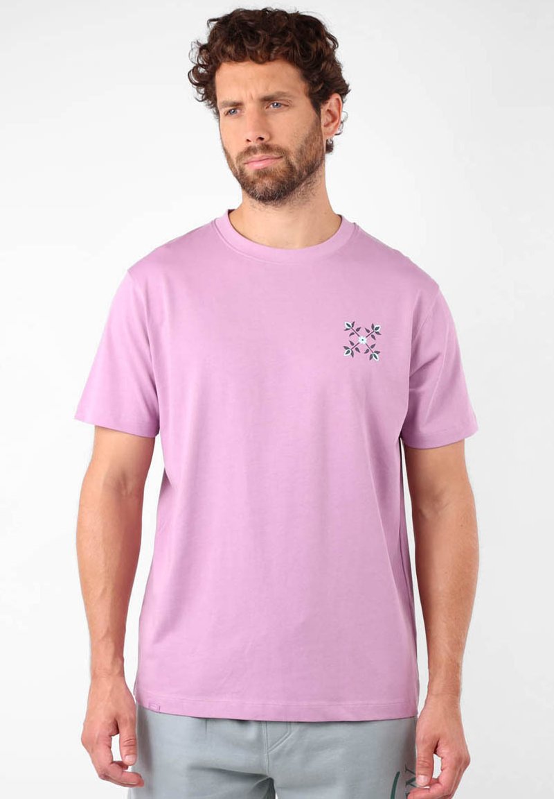 T-shirt en coton rose clair avec un col rond et des manches courtes. Présente un petit design décoratif avec des feuilles vertes sur la poitrine.