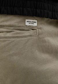 Beige cotton fabric with a stitched pocket, featuring a white label with black text "JACK & JONES EST. 1990" on the upper side.