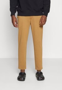 Beige Jogginghose mit entspannter Passform, ausgestattet mit einer Fronttasche, kombiniert mit schwarzen perforierten Clogs. Der Stoff wirkt weich und strukturiert.