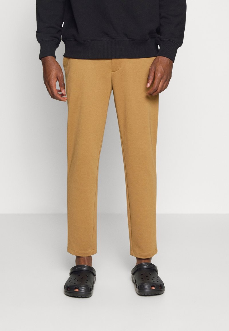 Beige Jogginghose mit entspannter Passform, ausgestattet mit einer Fronttasche, kombiniert mit schwarzen perforierten Clogs. Der Stoff wirkt weich und strukturiert.