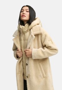 Cappotto in pelliccia finta crema con una texture morbida, grande colletto e chiusura a bottoni, indossato sopra un giubbotto imbottito beige chiaro con un collo alto.