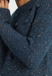 Pull tricoté bleu foncé avec des éclats multicolores. Il présente un col arrondi et des accents texturés en forme de boules sur l'épaule et le poignet.