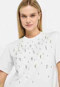 T-shirt bianco a maniche corte con decorazioni di perle e strass sparse sulla parte anteriore, che conferiscono un look texturizzato e impreziosito.