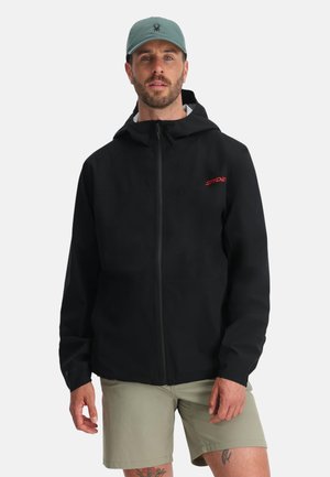 Mann in schwarzer Kapuzenjacke mit rotem Logo, beigen Shorts und grüner Mütze mit kleinem schwarzem Spinnenemblem.