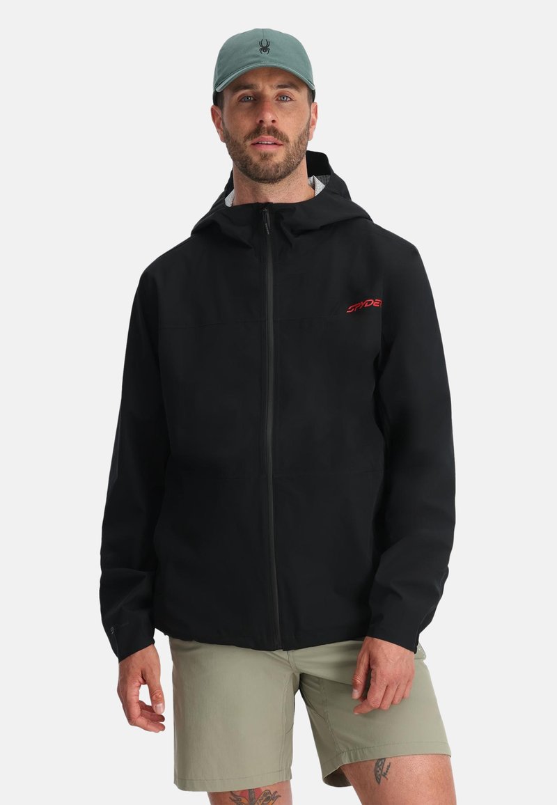 Uomo che indossa una giacca nera con cappuccio e logo rosso, pantaloni corti beige e un cappellino verde con un piccolo emblema di ragno nero.