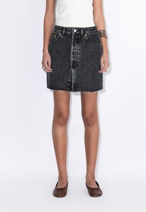 Vrouw draagt een zwarte denim minirok met hoge taille en rafelige zoom, een witte mouwloze top, bruine platte schoenen, en zilveren ringen en armband.