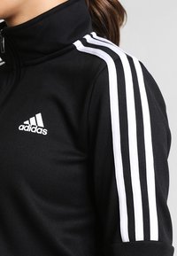 Gros plan d'une personne portant une veste de survêtement noire Adidas avec trois bandes blanches sur l'épaule et le logo Adidas sur la poitrine.