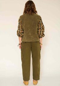 Un gilet vert texturé sur une chemise à carreaux. Un pantalon cargo olive avec des poches latérales. Des sabots en bois. Vue arrière, cheveux coiffés de manière lâche. Fond neutre.