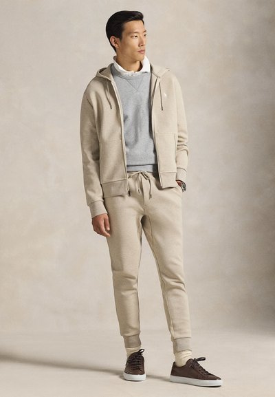 Muž nosiaci béžovú mikinu na zips, zodpovedajúce tepláky, sivú mikinu, bielu košeľu s golierom a tmavohnedé tenisky, stojaci s jednou rukou v vrecku.