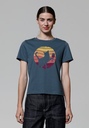 T-shirt en coton teal avec un graphique rond représentant un chat noir et un verre de vin, sur fond de design dégradé de coucher de soleil passant du jaune au rouge.