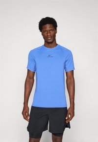 Homme athlétique debout, portant un t-shirt bleu Nike Pro à manches courtes et un short noir à plusieurs couches, faisant face à l'avant sur un fond uni.