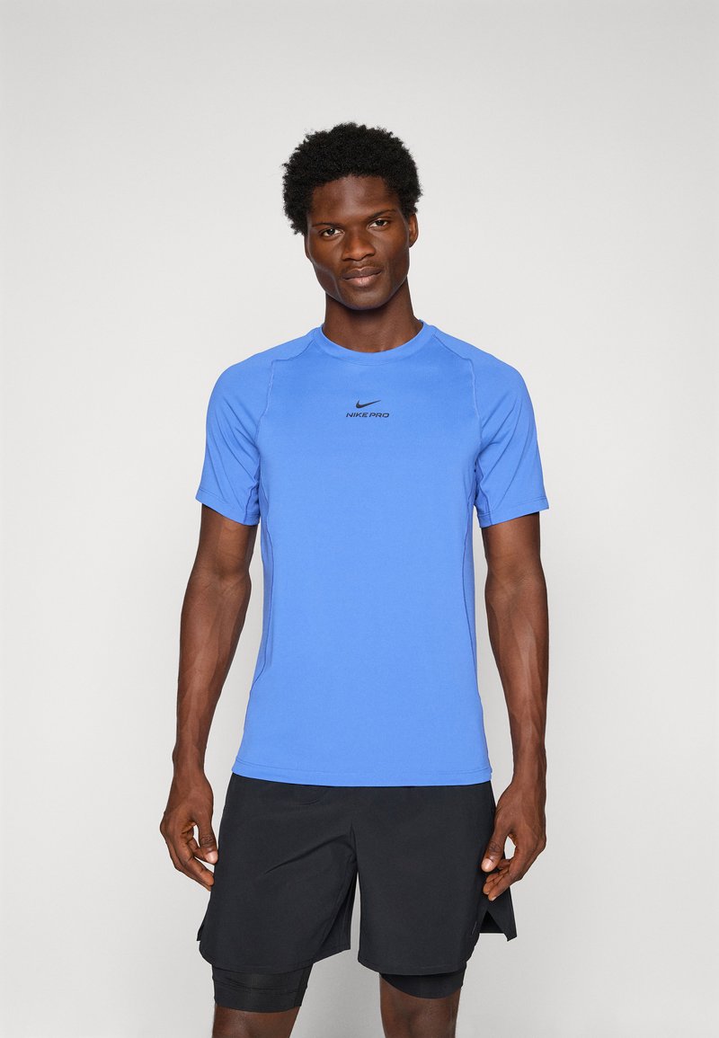 Homme athlétique debout, portant un t-shirt bleu Nike Pro à manches courtes et un short noir à plusieurs couches, faisant face à l'avant sur un fond uni.
