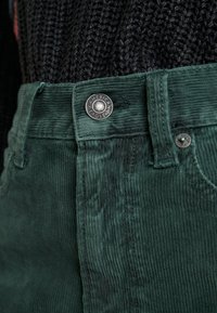 Gröna kordfläskbyxor med en metallknapp med "American Eagle"-logotyp, som visar en vertikal ribbad struktur och en skräddarsydd passform.