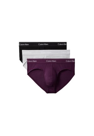 Jonge gespierde man staat blootsvoets, draagt alleen lichtgekleurde slip, tegen een effen grijze achtergrond.