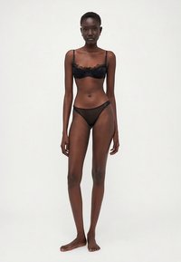 Črni čipkasti modrček brez košaric s tankimi naramnicami in usklajene prosojne črne tangice, okrašene z zapletenim robom in minimalističnim dizajnom. Gladek tekstilen material.