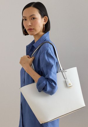 Γυναίκα με μπλε πουκάμισο που κρατάει μια μεγάλη λευκή δερμάτινη τσάντα ώμου με μαύρες λαβές πάνω στον ώμο, μπροστά σε απλό φόντο.