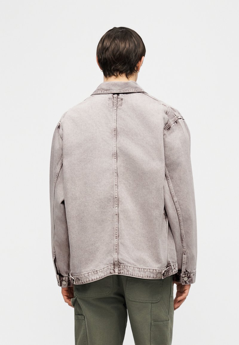 Personne aux cheveux courts portant une veste en denim gris clair et un pantalon vert, vue de dos sur un fond blanc uni.