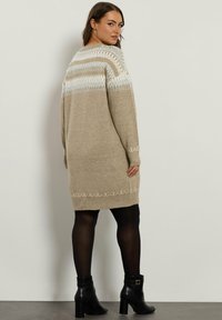 Robe pull en maille beige avec un motif géométrique blanc aux épaules, des poignets côtelés et un ourlet. Portée avec des bottines noires.