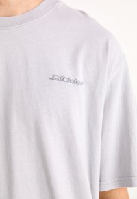 T-shirt de algodão cinza claro com mangas curtas, apresentando um pequeno logótipo bordado que diz "Dickies" na área do peito esquerdo.