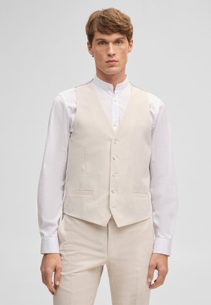 Junger Mann, der eine beigefarbene, geknöpfte Weste über einem weißen Langarmhemd und passende beigefarbene Hose trägt, steht vor einem schlichten Hintergrund.