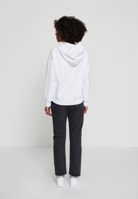 Vit huvtröja med ribbad mudd och nederkant, kombinerad med mörkgrå raka jeans och vita sneakers. Enfärgad bakgrund.