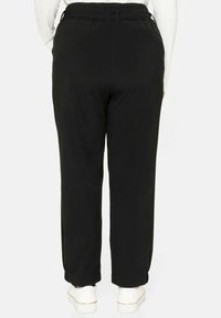 Zadní pohled na osobu v černých kalhotách rovného střihu s elastickým pasem, v kombinaci s bílými teniskami a zasunutou bílou košilí.