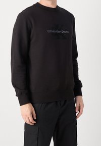 Czarna bluza wykonana z miękkiego materiału, z okrągłym dekoltem, ściągaczami przy mankietach i u dołu, z logo "Calvin Klein Jeans" na środku.