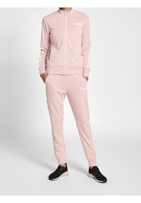 Rosa träningsdräkt i mjukt tyg, med en zip-up jacka och matchande byxor med vitt logo och chevrondetaljer på ärmar och sidor. Svarta sneakers.