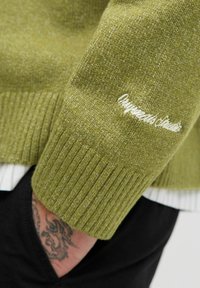 Pogled od blizu na zeleni pleteni jumper z belim vezenjem "Concrete Studio" nad manšeto ter tetovirano roko v črni žep.