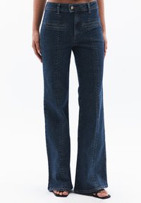 Jeans de mezclilla oscuros de talle alto y acampanados con bolsillos frontales y costuras a tono. Presentan un cierre de botón clásico y un corte relajado en la pierna.