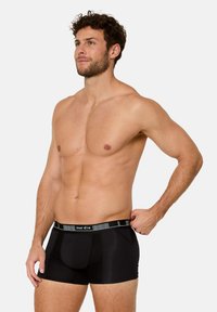 Skjortlös man med lockigt hår som bär svarta Nur Die-boxershorts, står med ena handen och justerar midjebandet, tittar åt vänster, neutral bakgrund.