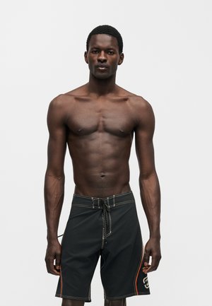 Athletischer Mann, der frontal steht, trägt schwarze Badeshorts mit orangefarbenen Akzenten vor einfachem hellem Hintergrund.