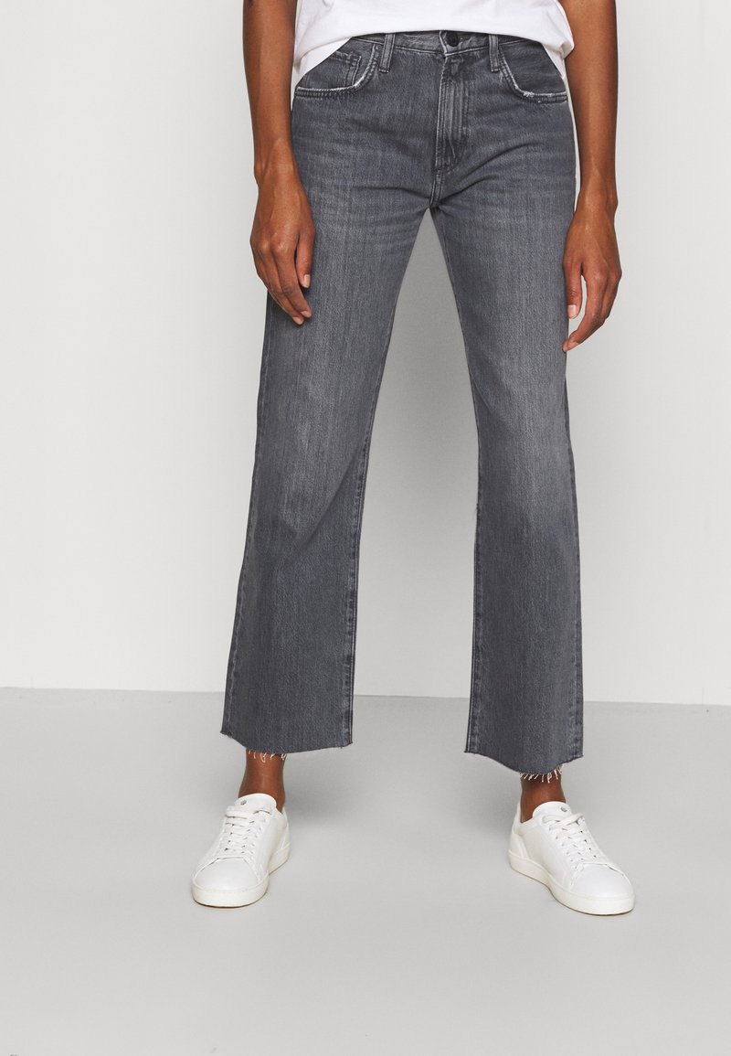 Jeans grigi ampi con orlo grezzo, design a cinque tasche e vita aderente, abbinati a sneaker bianche su uno sfondo neutro.