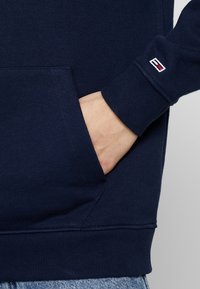 Mano en el bolsillo de un sudadera azul marino con un pequeño logo en el puño, combinada con unos jeans de mezclilla de color azul claro.