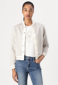 Texturierte Bomberjacke in Elfenbein und Gold, mit geometrischem Muster, gerippten Bündchen und Knopfschluss. Über einem weißen Oberteil getragen.