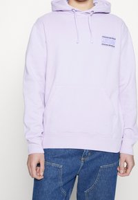 Ljust lila hoodie i bomullsblandning, med en framficka, dragsko på huvan och en liten grafisk patch på bröstet.