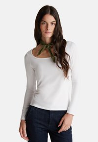 Donna con lunghi capelli scuri che indossa una maglia bianca a maniche lunghe, jeans scuri e una sciarpa verde oliva su uno sfondo chiaro e semplice.