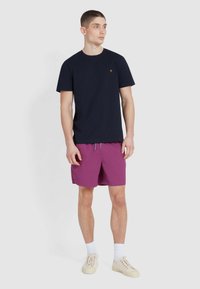 Navy blauw t-shirt met korte mouwen en een klein geel logo, gecombineerd met paarse shorts. Model draagt lichte sneakers en witte sokken.