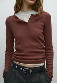 Roestrood langetermijnshirt met een henleyhalslijn, drie knopen, gedragen over een witte gelaagde blouse, gecombineerd met hoge grijze broeken en een zwarte riem.