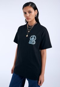 Camiseta de algodón negra con un gráfico abstracto azul en el lado izquierdo del pecho. Corte holgado con mangas cortas, cuello redondo y superficie texturizada.