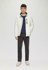 Leichte Steppjacke in Off-White mit hohem Kragen und Reißverschluss vorne; kombiniert mit einem marineblauen Pullover und dunklen Hosen, dazu weiße Sneaker.