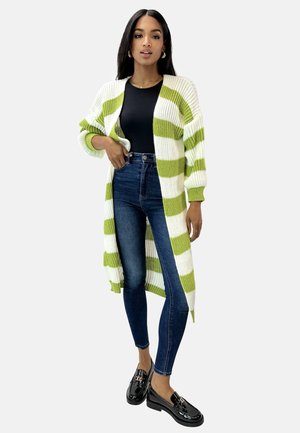 Kvinde iført lang hvid og grønstribet cardigan, sort top, blå tætsiddende jeans og sorte loafers, stående mod en ensfarvet baggrund.