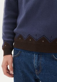 Virkad tröja i marinblått med en zigzag-brun fåll och ljusblå accenter. Har ribbade ärmslut och detaljerad sömnad vid fållen.