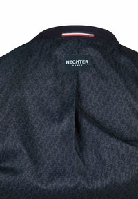 Wnętrze ciemnej kurtki z powtarzającym się wzorem H, czarnym kołnierzem z paskiem w kolorach czerwonym, białym i niebieskim oraz prostokątną etykietą HECHTER PARIS na środku.