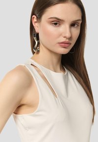Beżowa bluzka bez rękawów z wycięciami na ramionach. Modelka nosi duże, skręcone srebrne kolczyki. Gładki materiał, minimalistyczny design.