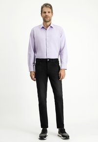Camicia leggera viola con taschino sul petto, abbinata a jeans neri. Il modello indossa sneaker scure, in piedi contro uno sfondo semplice.