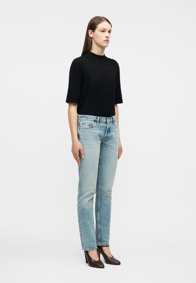 Γυναίκα με μαύρο ριμπ μπλουζάκι με κοντά  μανίκια, ανοιχτό μπλε τζιν παντελόνι με ίσια γραμμή και καφέ ψηλοτάκουνα παπούτσια, στέκεται μπροστά σε λευκό απλό φόντο.