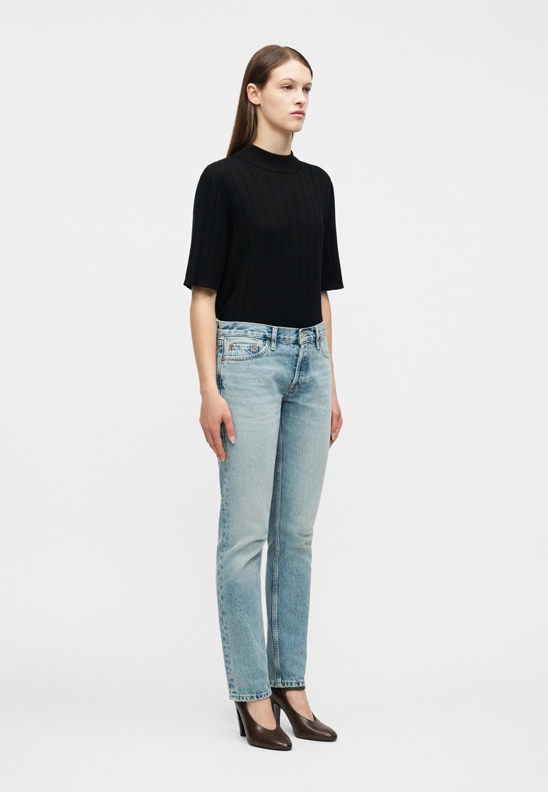 Femme portant un haut noir à manches courtes à côtes, un jean bleu clair droit et des chaussures à talons hauts marron, debout devant un fond blanc uni.