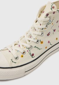 Högskor av canvas i krämfärg med flerfärgad blommönsterbroderi, platta vita snören och en gummitåhätta; svarta och vita sula accenter.